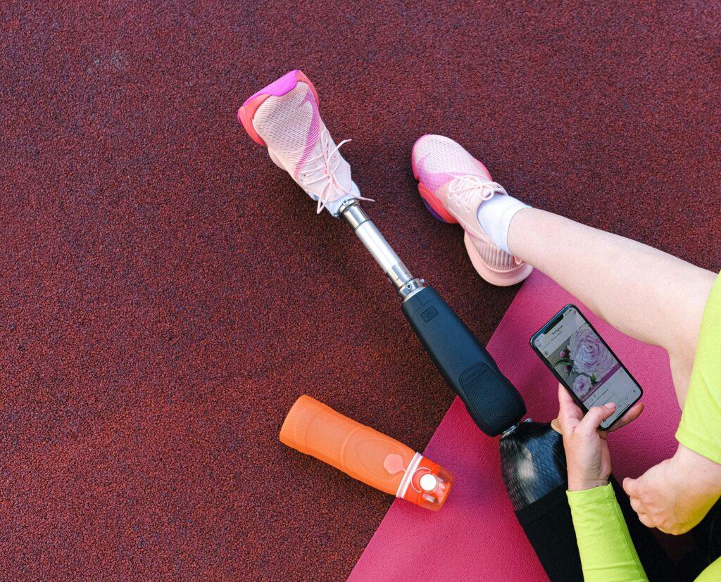 A woman with a prosthetic leg sits on a yoga mat, using her smartphone in casual sportswear.