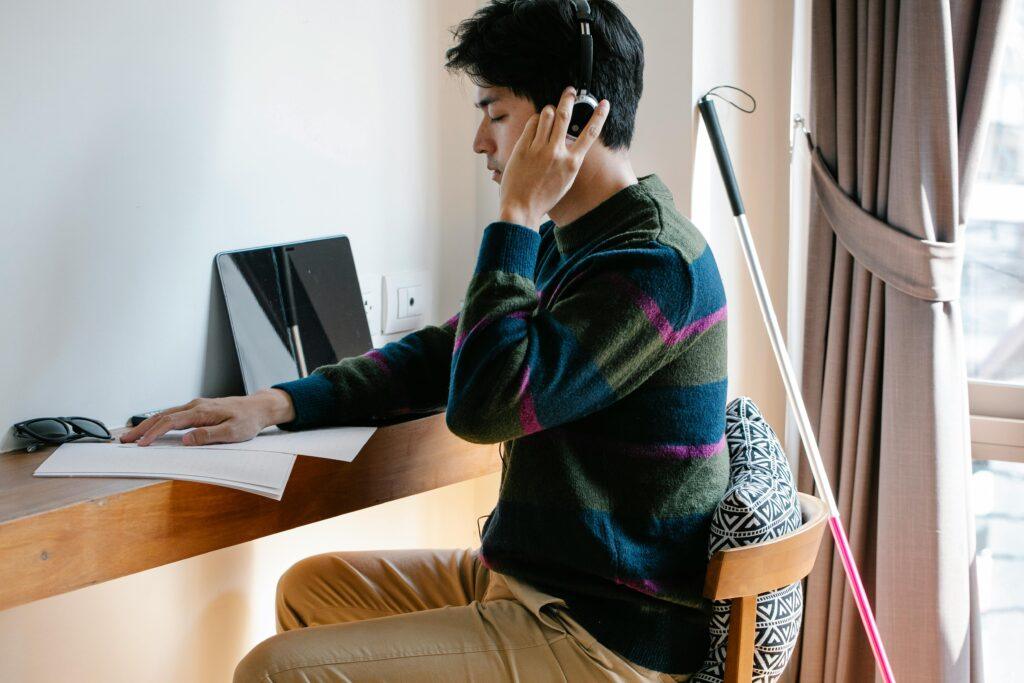 A visually impaired man uses headphones and reads braille indoors at a wooden desk.